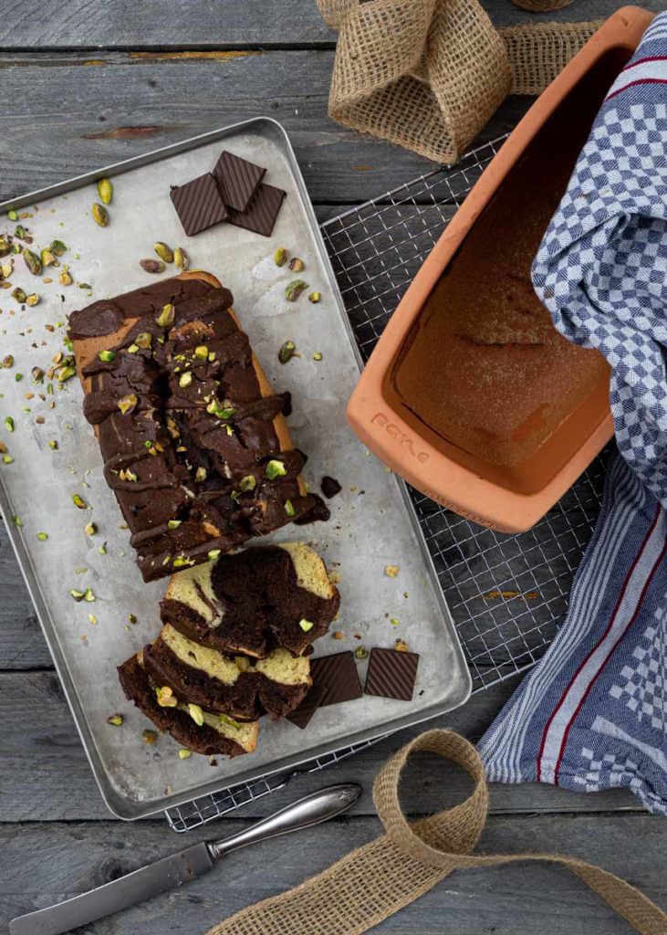 Schokoladen-Pistazien-Kuchen auf einem Blech mit Schokostücken, geschnitten, neben leere Backform und Tuch.