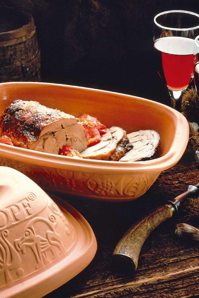 Bräter mit gebratenem Fleisch und Gemüse, daneben Wein, Deckel und Messer auf rustikalem Holztisch.