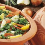 Keramiktopf mit Gemüseeintopf aus Erbsen, Karotten und Kartoffeln. Daneben Zwiebeln, Kohlrabi und Petersilie.