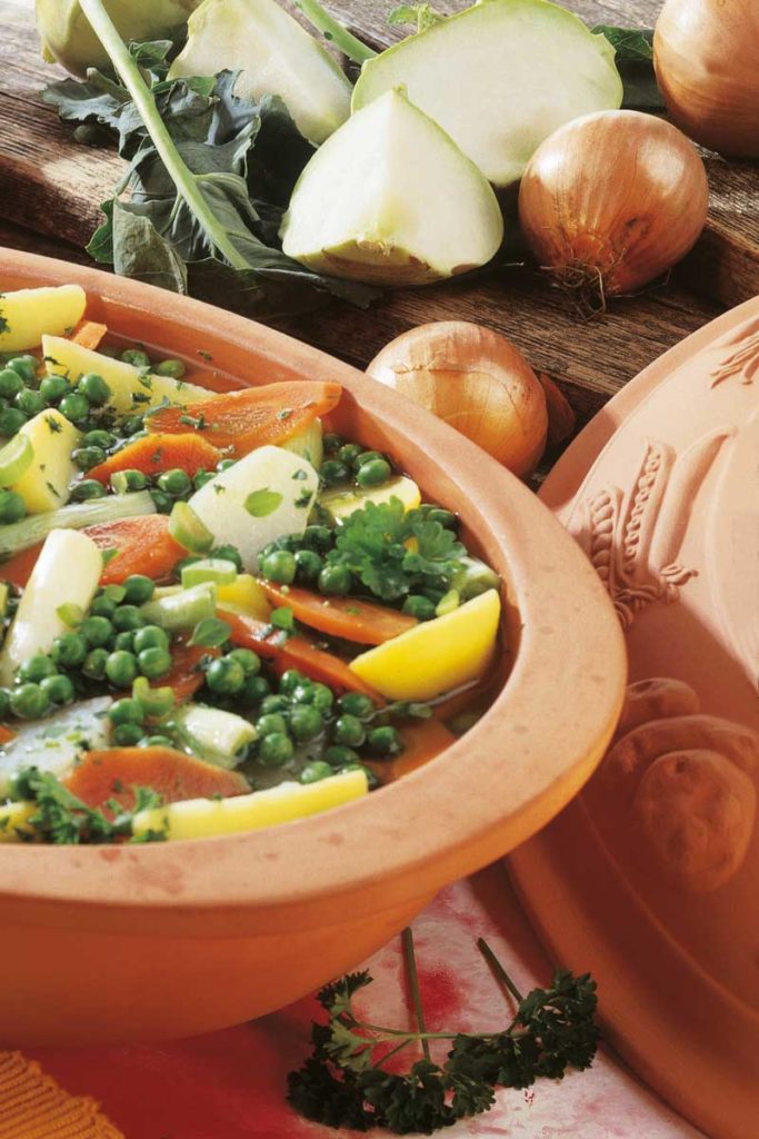 Keramiktopf mit Gemüseeintopf aus Erbsen, Karotten und Kartoffeln. Daneben Zwiebeln, Kohlrabi und Petersilie.