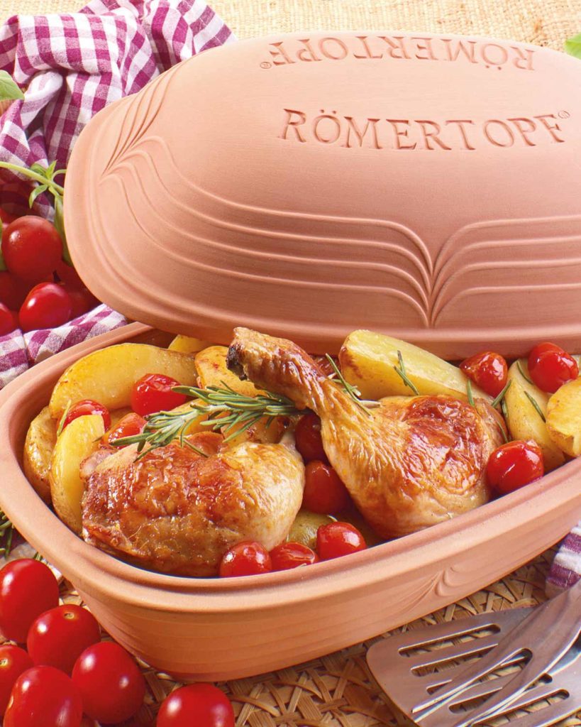 Brathähnchenkeulen mit Kartoffeln und Tomaten im Römertopf, dekoriert mit Rosmarin, auf einem karierten Tuch.