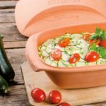 Terrakotta-Römertopf mit Zucchini und Tomaten auf Holztisch, daneben frische Tomaten und Zucchini.