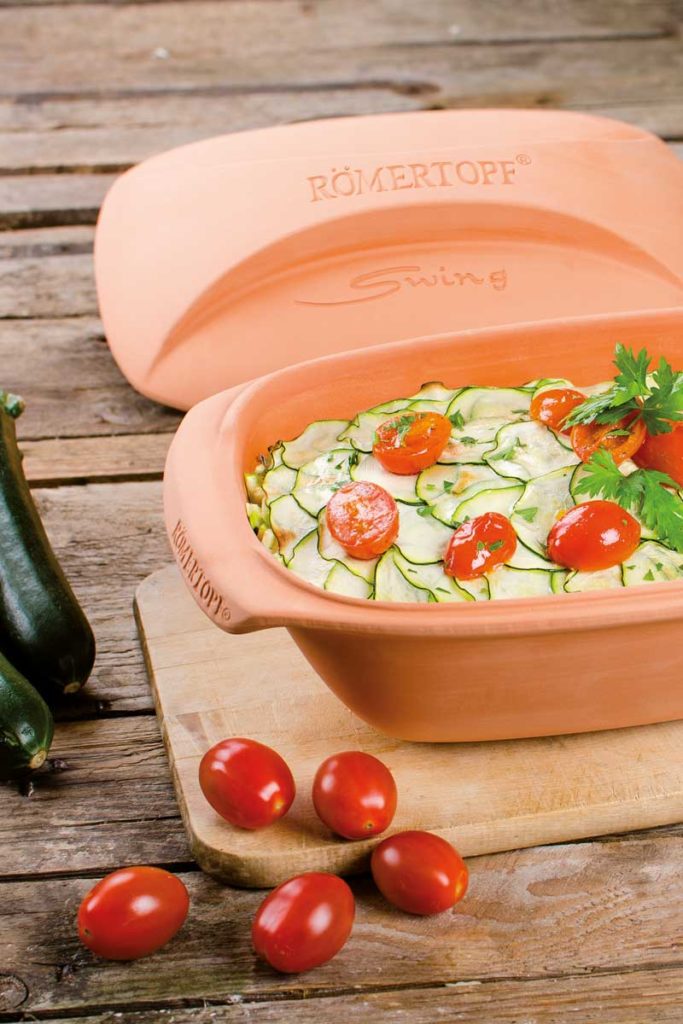 Terrakotta-Römertopf mit Zucchini und Tomaten auf Holztisch, daneben frische Tomaten und Zucchini.