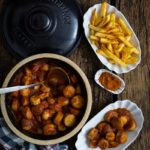 Ein Topf mit Currywurst, beigelegte Pommes, Gewürz und eine Serviette auf rustikalem Holztisch.