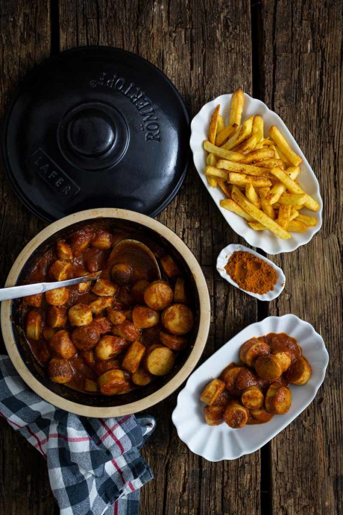 Ein Topf mit Currywurst, beigelegte Pommes, Gewürz und eine Serviette auf rustikalem Holztisch.