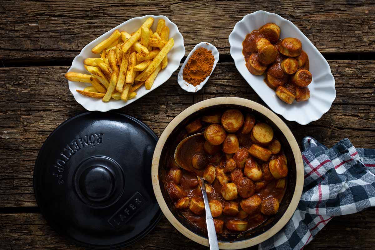 Schüssel mit Currywurst in Soße, daneben Schalen mit Pommes und Currypulver, auf einem Holztisch.