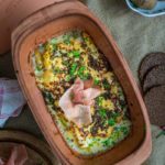 Überbackener Auflauf im römischen Tontopf mit Schinken und frischen Kräutern, begleitet von Brot und Kartoffeln.