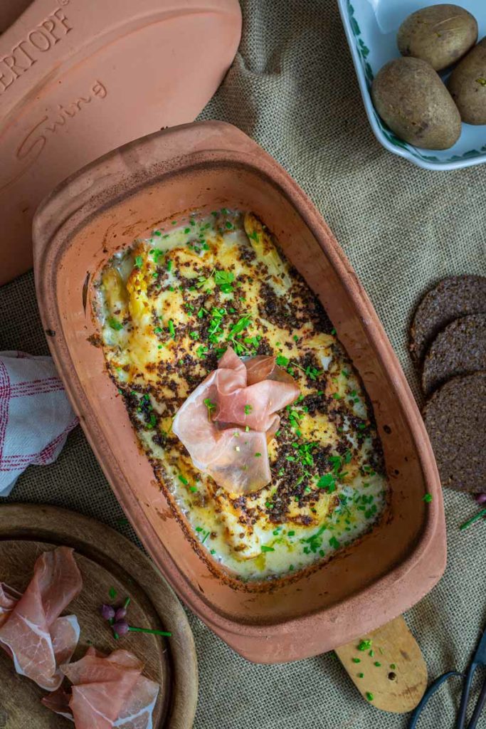 Überbackener Auflauf im römischen Tontopf mit Schinken und frischen Kräutern, begleitet von Brot und Kartoffeln.