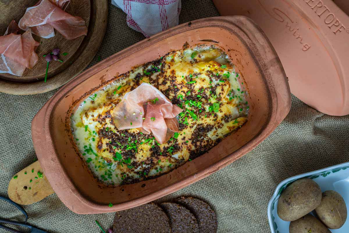 Auflauf in Keramikform mit Kräutern und Schinken garniert, daneben Brot und rohe Kartoffeln.