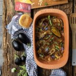 Auberginen-Auflauf im Römertopf, daneben Käse, Gewürze, frische Auberginen und ein Küchentuch auf einem Holztisch.