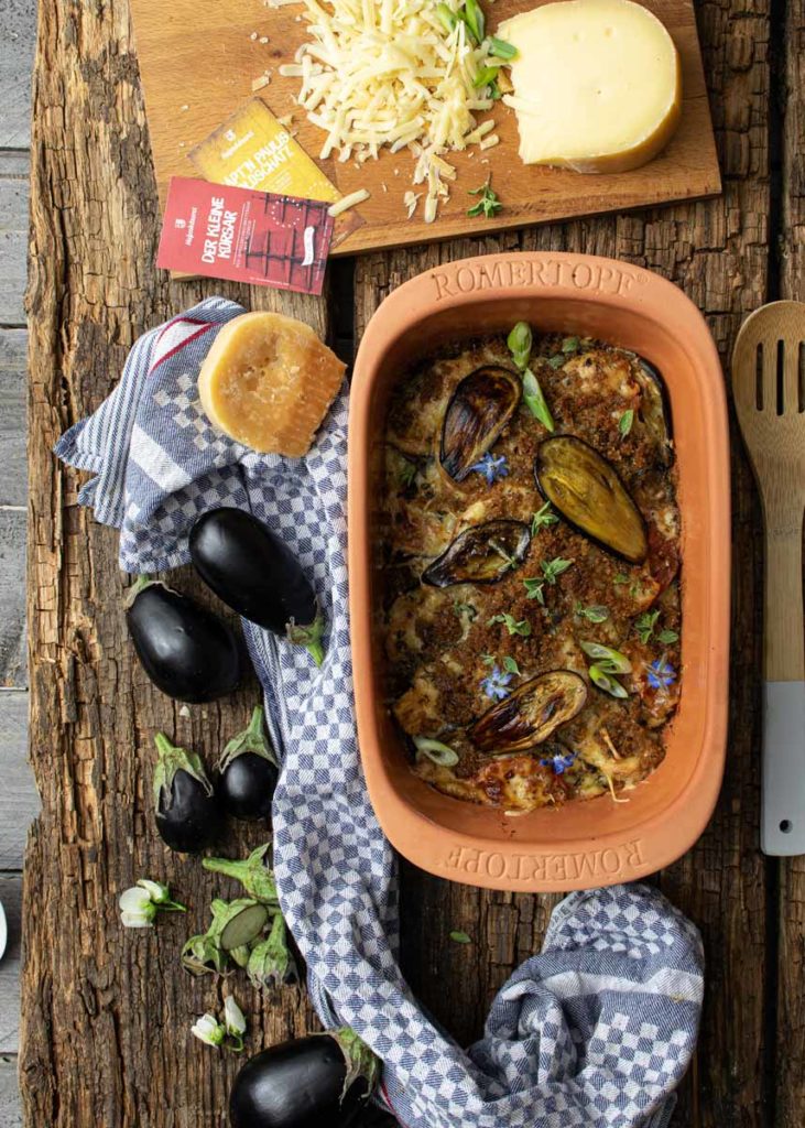 Auberginen-Auflauf im Römertopf, daneben Käse, Gewürze, frische Auberginen und ein Küchentuch auf einem Holztisch.