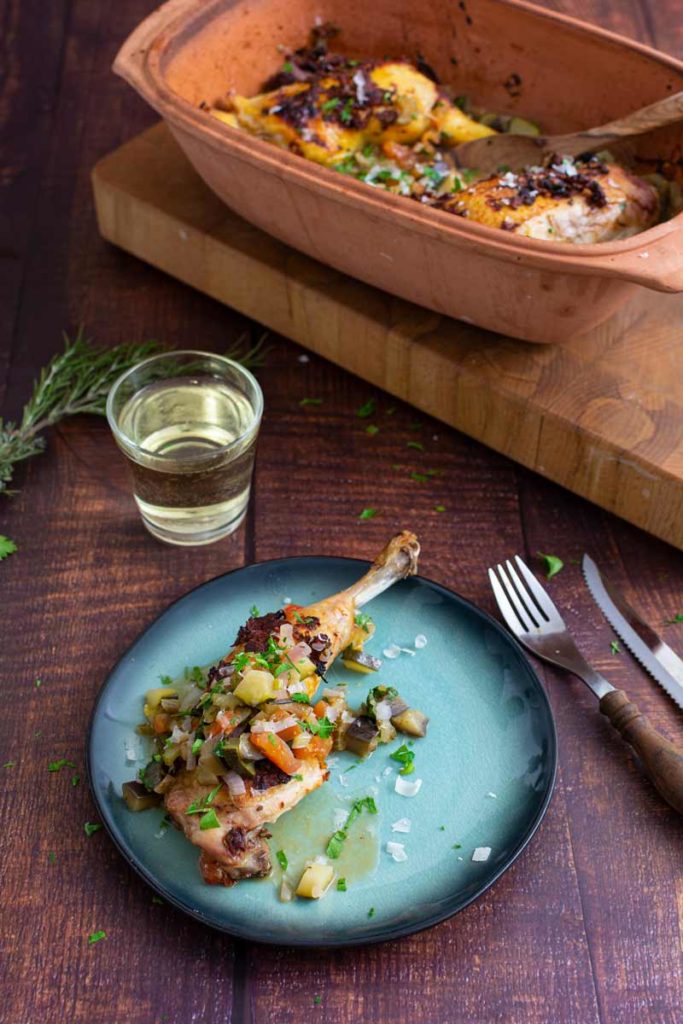 Hähnchenkeule mit Gemüse auf einem Teller, daneben ein Glas Weißwein und eine Auflaufform im Hintergrund.