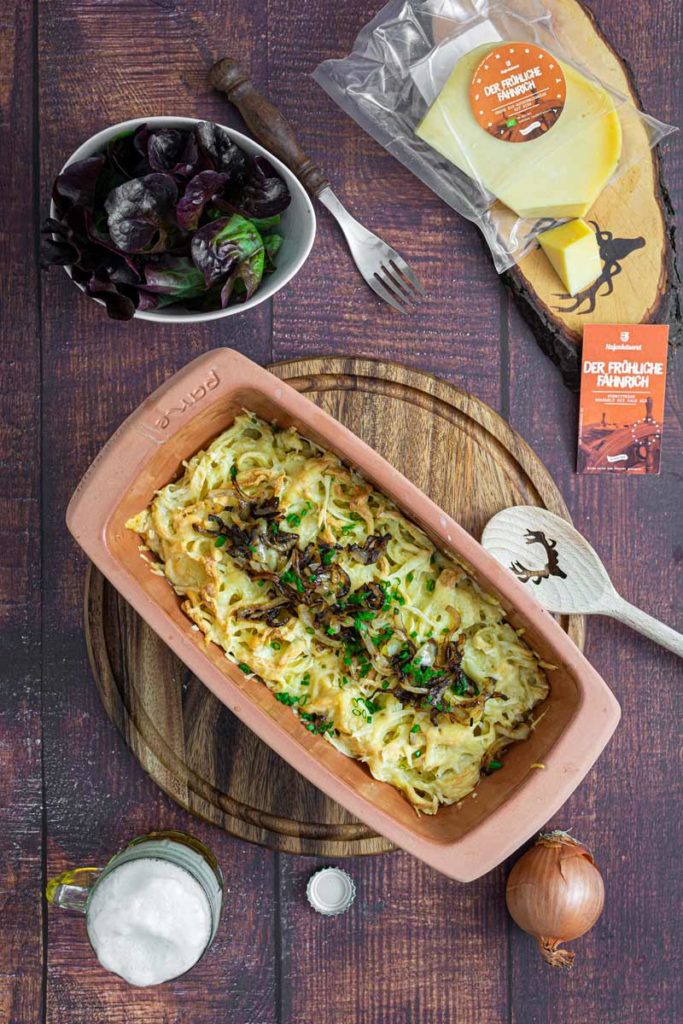Auflaufform mit Kässpätzle und Röstzwiebeln, daneben Salat, Käse, ein Bier und eine Zwiebel auf einem Holztisch.