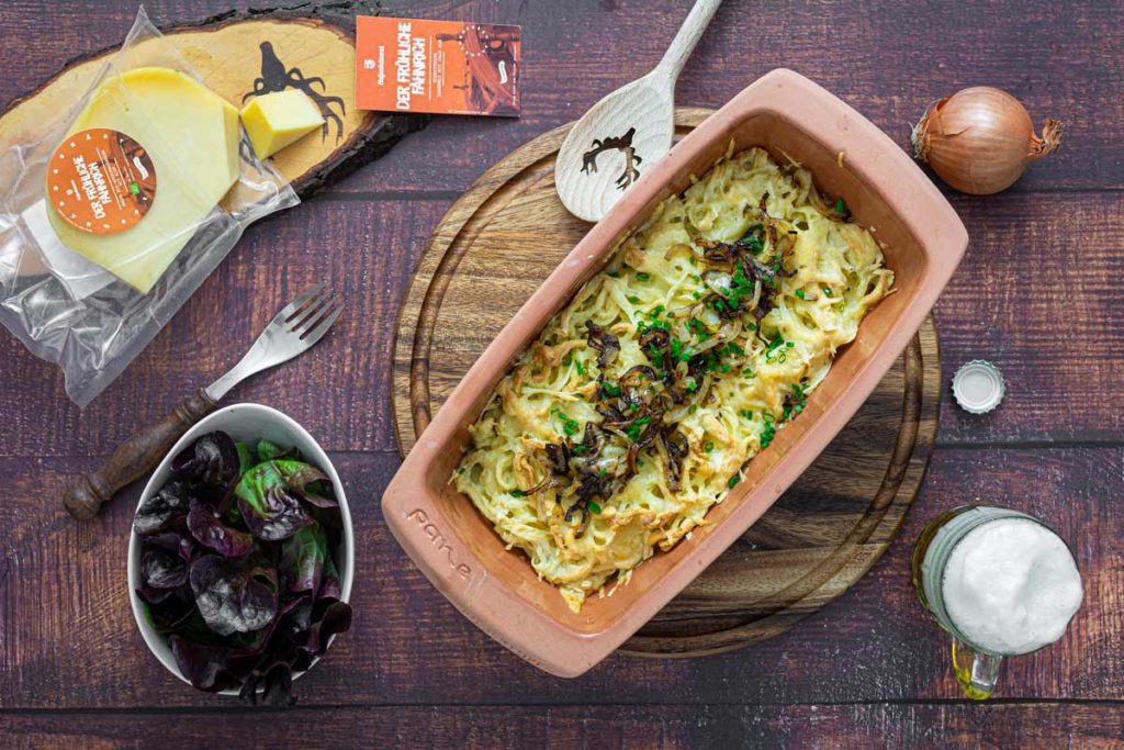Auflaufform mit Käsespätzle, daneben Käsescheiben, Salat, Zwiebel und Bierkrug auf Holztisch.