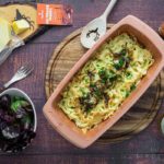 Auflaufform mit Käsespätzle, daneben Käsescheiben, Salat, Zwiebel und Bierkrug auf Holztisch.