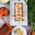Karottenkuchen mit Frischkäseglasur und Dekoration; umgeben von Karotten, Nüssen und Kochutensilien auf Holztisch.