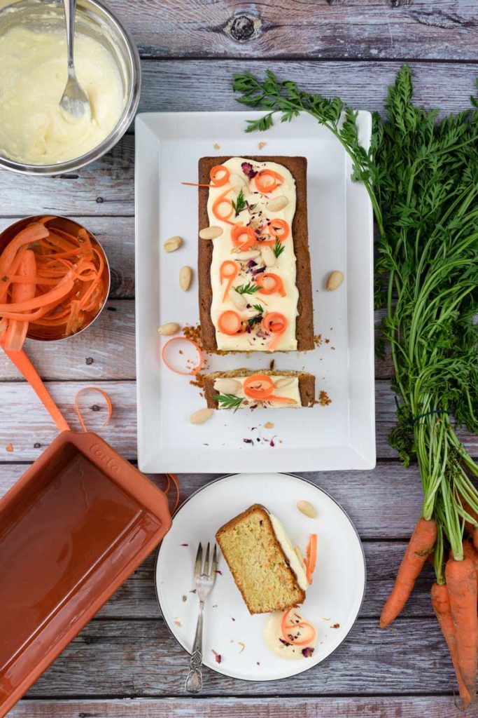 Karottenkuchen mit Frischkäseglasur und Dekoration; umgeben von Karotten, Nüssen und Kochutensilien auf Holztisch.