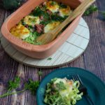 Nudelauflauf mit Brokkoli und Schinken in einer Terrakotta-Form, daneben ein Teller mit einer Portion, auf Holztisch.