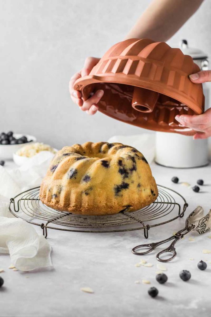 Hand hebt Gugelhupfform von einem frisch gebackenen Heidelbeerkuchen auf einem Kuchengitter.