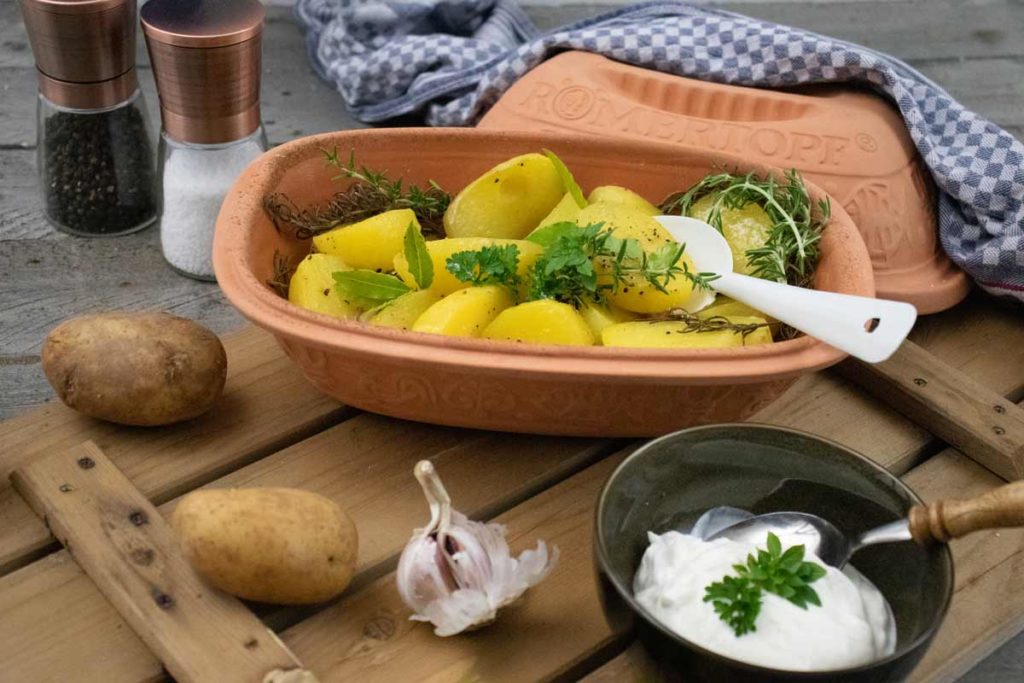 Römertopf mit Kartoffeln und Kräutern, daneben Schälchen mit Dip, Knoblauchknolle und Gewürzstreuer auf Holzbrett.