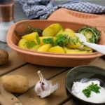 Römertopf mit Kartoffeln und Kräutern, daneben Schälchen mit Dip, Knoblauchknolle und Gewürzstreuer auf Holzbrett.