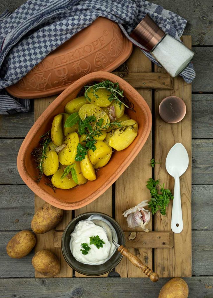 Ofen-Kartoffeln mit Kräutern in einem Tontopf, daneben Joghurt-Dip, Kräuter und Knoblauch auf Holzbrett.