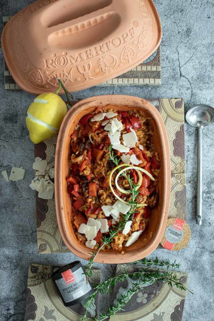 Römertopf mit Gemüseauflauf, garniert mit Käse und Kräutern. Daneben eine Zitrone, Kräuterzweig und Pfeffer.