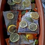 Roher Fisch in einem Bräter, belegt mit Zitronenscheiben, Lorbeerblättern und Pfeffer, auf Gemüse wie Karotten.