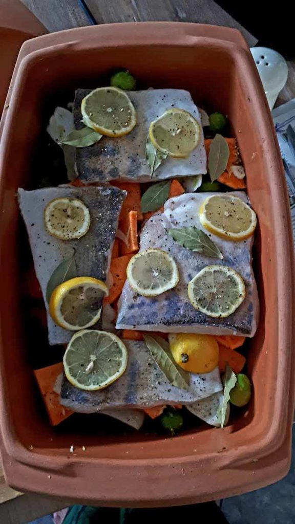 Roher Fisch in einem Bräter, belegt mit Zitronenscheiben, Lorbeerblättern und Pfeffer, auf Gemüse wie Karotten.