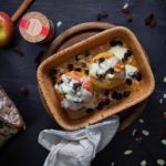 Gebackene Äpfel mit Rosinen, Mandelblättchen und Vanillesoße in einer Keramikform auf dunklem Untergrund.