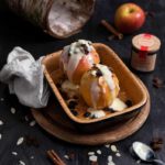 Zwei gebackene Äpfel mit Soße, Rosinen und Mandeln in einer Keramikform auf Holzteller, dekorativ mit Zimtstangen.