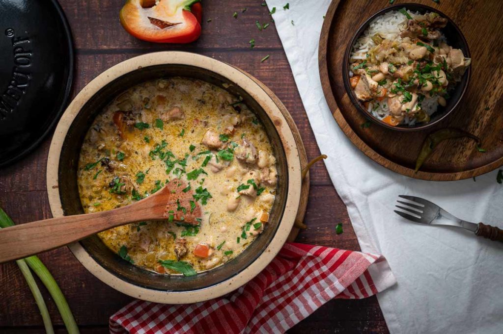 Eintopf in einem Topf mit Holzlöffel, dekoriert mit Petersilie. Serviert auf Reis in Schale. Paprika und Tuch daneben.