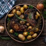 Gebratene Koteletts und Kartoffeln mit Rosmarin auf einem Holztisch, daneben Zwiebeln, Salz und ein Küchentuch.