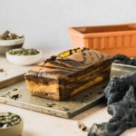 Marmorierter Kuchen auf einem Blech, mit Schokoladenstücken und Kürbiskernen, umgeben von Schüsseln und einer Backform.