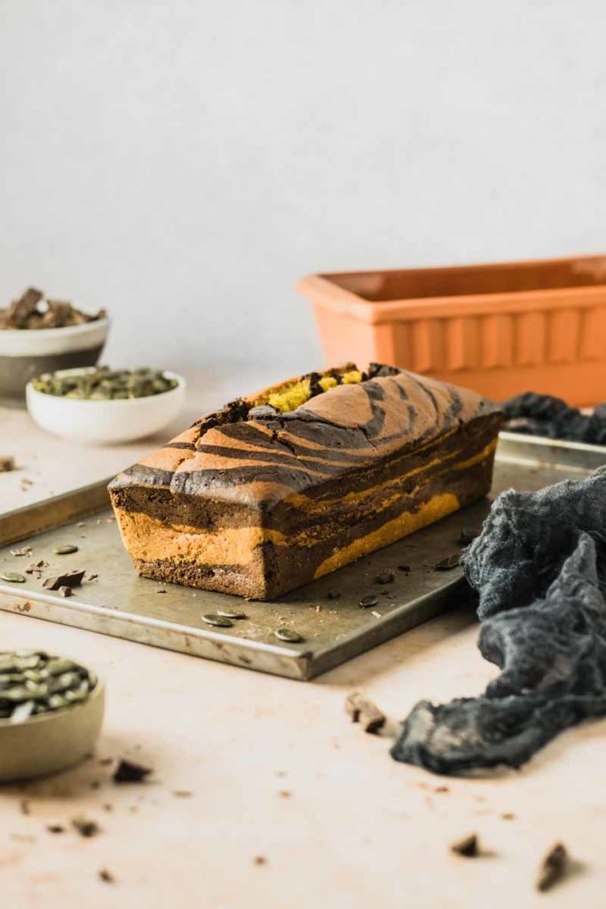 Marmorierter Kuchen auf einem Blech, mit Schokoladenstücken und Kürbiskernen, umgeben von Schüsseln und einer Backform.