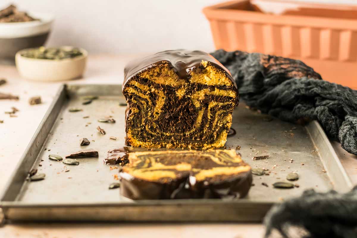 Ein angeschnittener Marmorkuchen mit Schokoladenglasur auf einem Blech, umgeben von Schokoladenstückchen und Kürbiskernen.