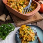 Kürbis-Casserole in einer braunen Auflaufform, serviert auf einem Teller mit Gabel, umgeben von Kürbissen und Petersilie.