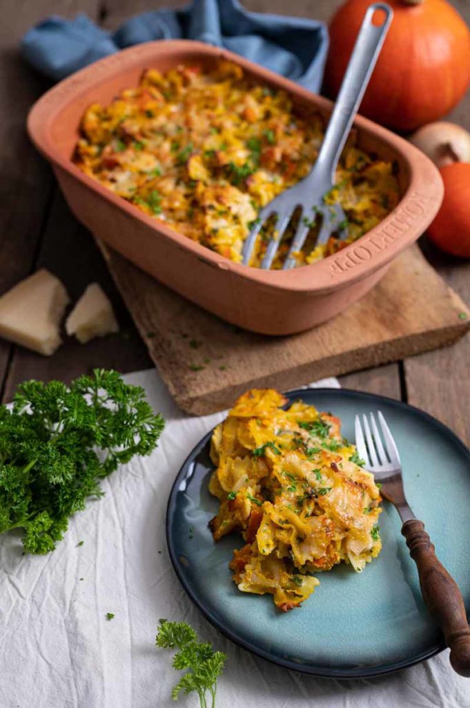 Kürbis-Casserole in einer braunen Auflaufform, serviert auf einem Teller mit Gabel, umgeben von Kürbissen und Petersilie.