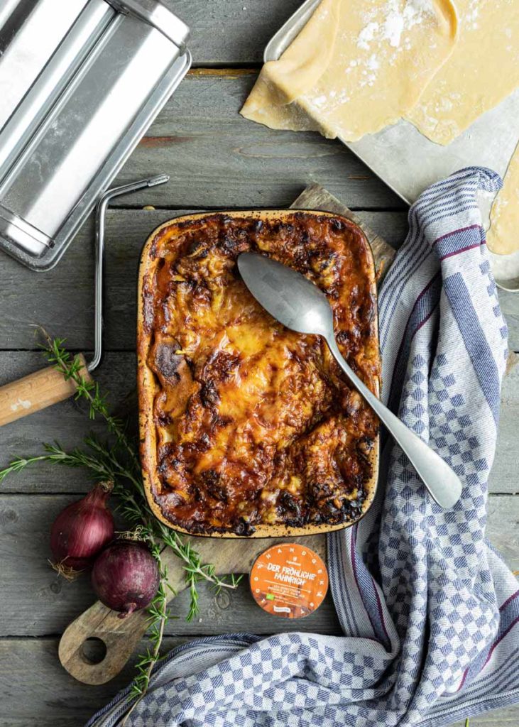 Lasagne in einer Auflaufform mit Löffel, Pizzateig, Zwiebeln und Kräutern auf Holztisch, neben Handtuch und Pastamaschine.