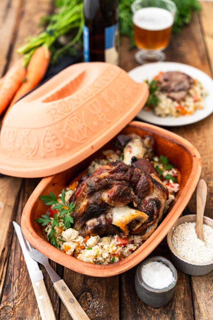 Schmorbraten mit Gemüse und Reis in einem Tonbräter auf Holztisch, daneben Salz, Quinoa und ein gedeckter Tisch.