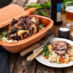 Tontopf mit Fleisch und Gemüse auf Couscous, angerichtet mit Besteck, Salz, Bierglas und Flasche auf Holztisch.