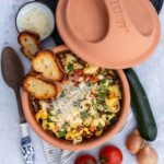 Terrakotta-Topf mit gefüllten Nudeln, geriebenem Käse und Gemüse, daneben Tomaten, Zucchini, Zwiebeln und geröstetes Brot.