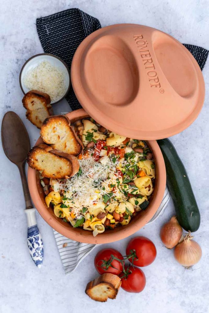 Terrakotta-Topf mit gefüllten Nudeln, geriebenem Käse und Gemüse, daneben Tomaten, Zucchini, Zwiebeln und geröstetes Brot.