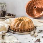 Gugelhupf auf Teller, umgeben von Schokolade, mit Gugelhupfform und Besteck im Hintergrund, auf grauem Untergrund.