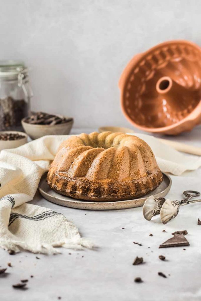 Gugelhupf auf Teller, umgeben von Schokolade, mit Gugelhupfform und Besteck im Hintergrund, auf grauem Untergrund.
