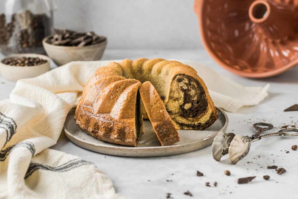 Ein Marmorkuchen auf einem Teller, zwei Stücke angeschnitten. Im Hintergrund eine Gugelhupfform und Schokoladenstücke.