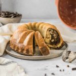 Ein Marmorkuchen auf einem Teller, zwei Stücke angeschnitten. Im Hintergrund eine Gugelhupfform und Schokoladenstücke.