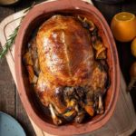 Gebratene Ente in einer Tonform, umgeben von Orangenstücken. Im Hintergrund Rotkohl und Soße.