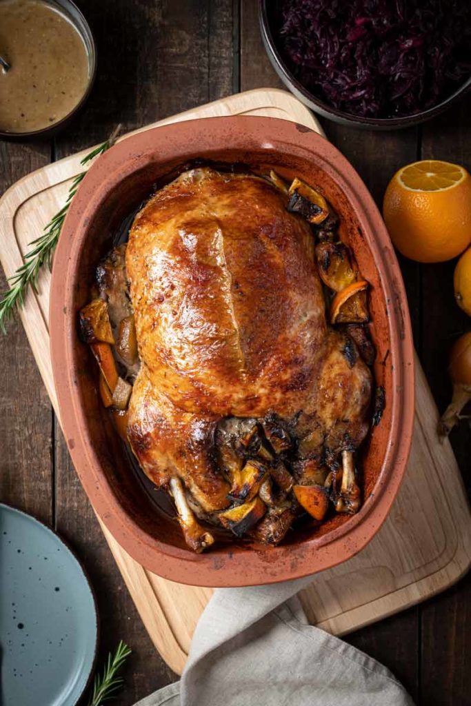 Gebratene Ente in einer Tonform, umgeben von Orangenstücken. Im Hintergrund Rotkohl und Soße.