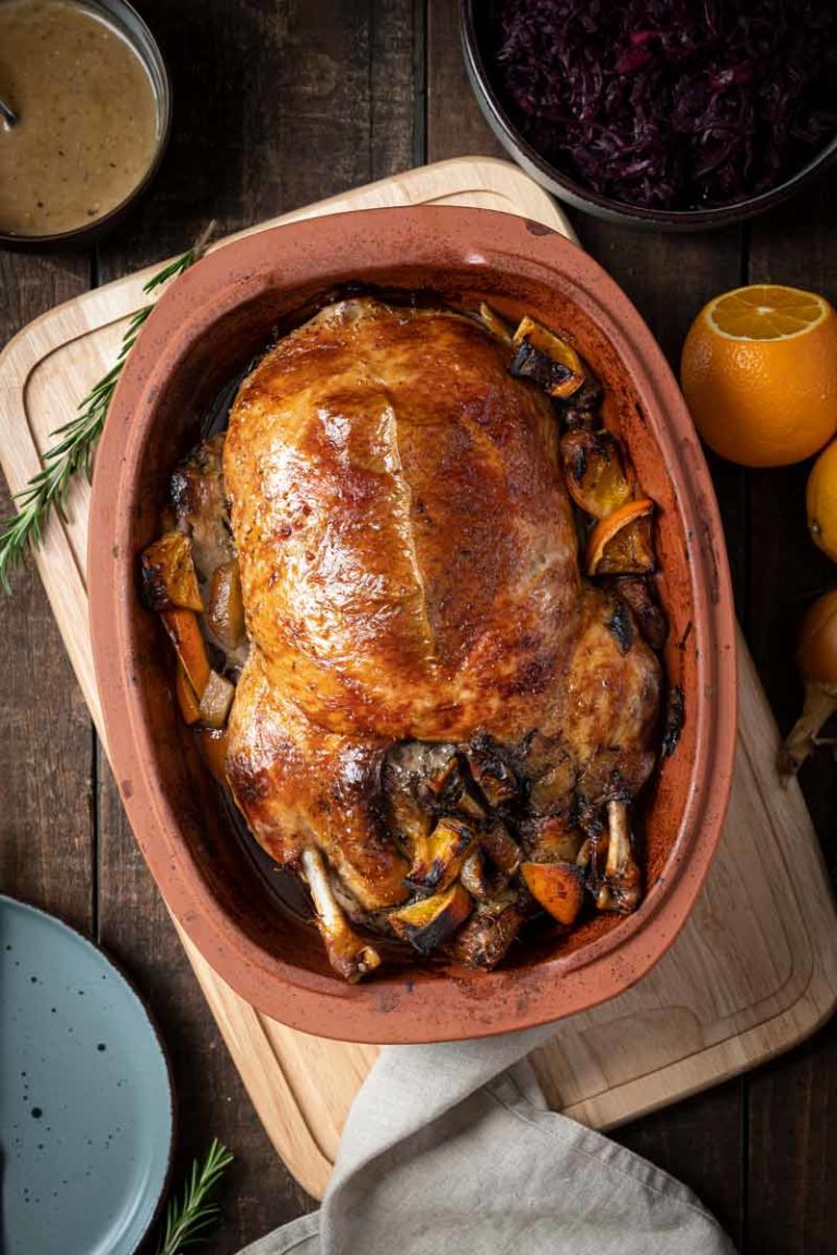 Gebratene Ente in einer Tonform, umgeben von Orangenstücken. Im Hintergrund Rotkohl und Soße.